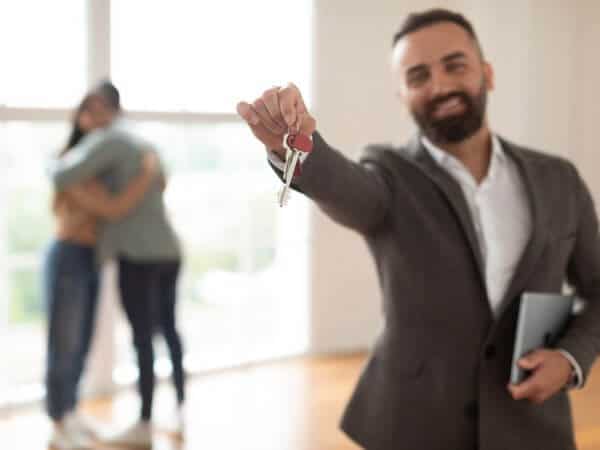 A real estate agent smiling and holding a key with a couple of tenants embracing each other in the background.