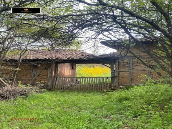 Village house from a plot for sale in the village of Ochusha, with green grass and trees around it. The house looks old with tiled roof and wooden fence.