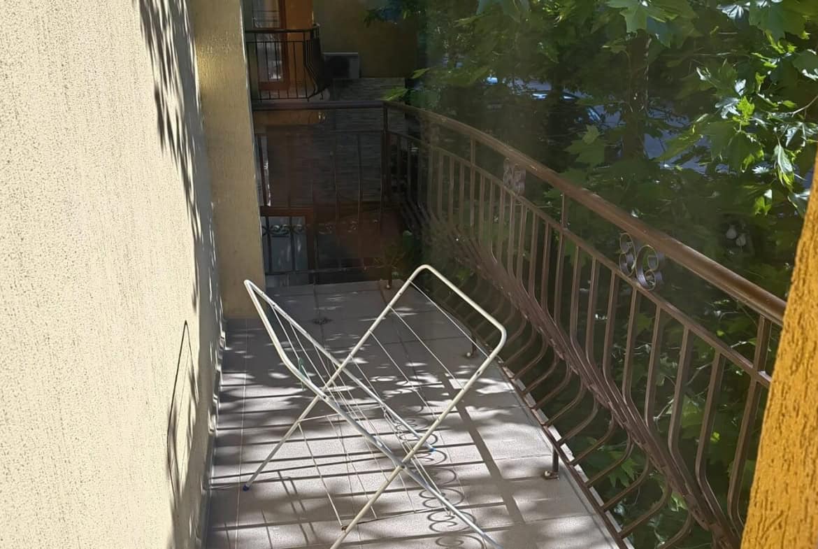 Балкон с метална сушилня за дрехи и зелени дървета на фона.