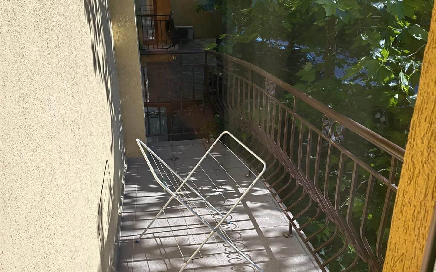 tristaen-apartament-prodajba-zlatni-pqsaci-1 Балкон с метална сушилня за дрехи и зелени дървета на фона.