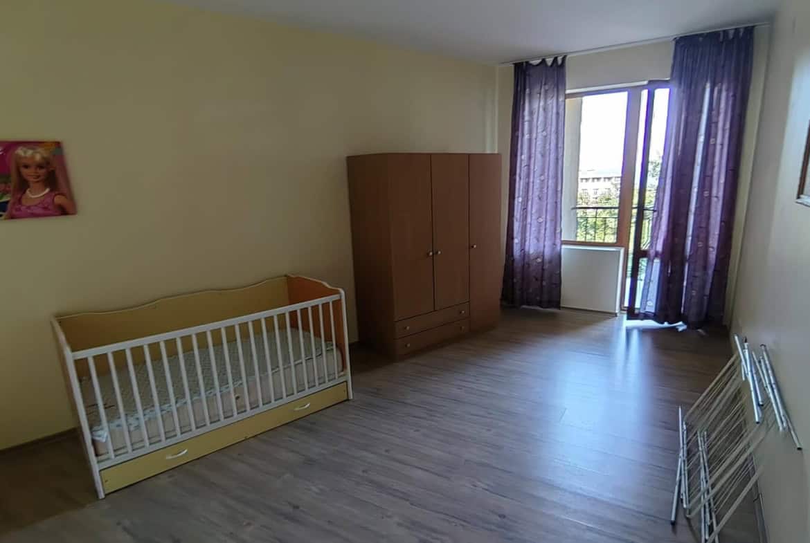 Спалня с бебешко креватче, голям кафяв гардероб и изглед към балкон.