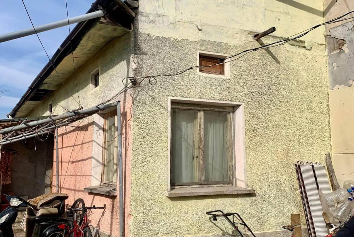 Външна фасада на къща с изкривен покрив и няколко прозореца, околната област е пълна с велосипеди и мотопед.