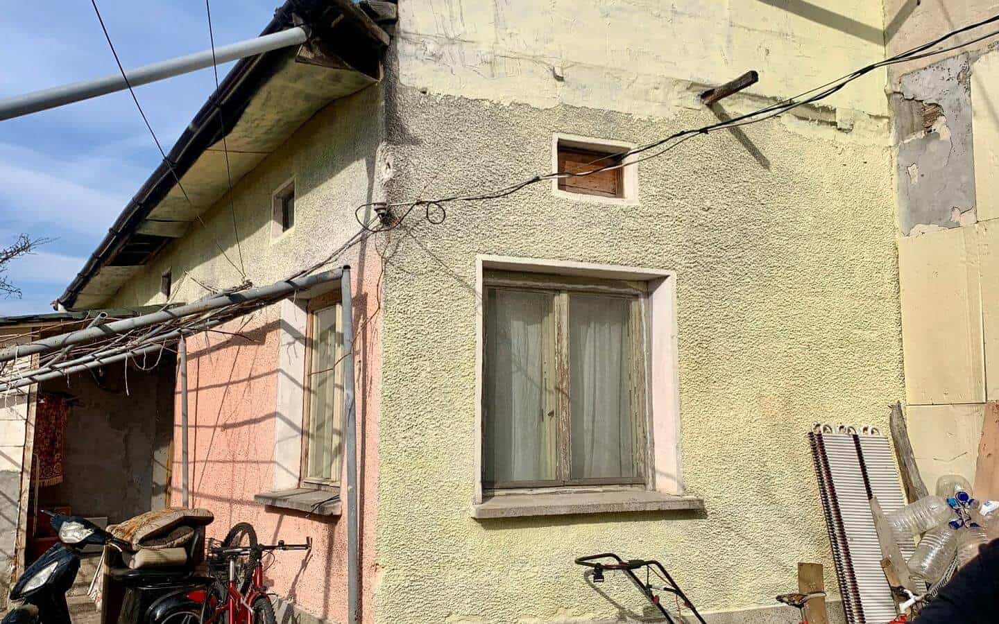 kushta-prodajba-kostinbrod-sofia-3 Външна фасада на къща с изкривен покрив и няколко прозореца, околната област е пълна с велосипеди и мотопед.
