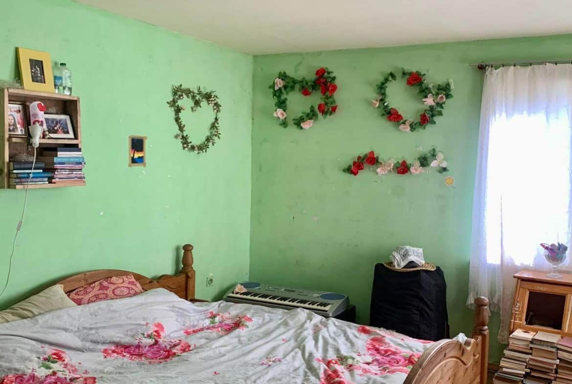 Спалня със зелени стени, декорирана с венци от изкуствени цветя и книги до леглото.