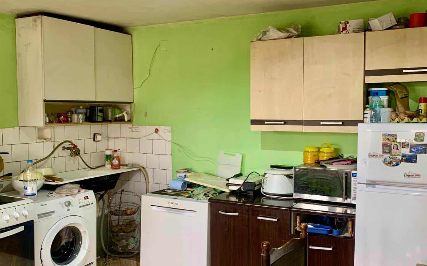 kushta-prodajba-kostinbrod-sofia-7 Кухня със светлозелени стени и различни кухненски уреди, включително пералня и хладилник.