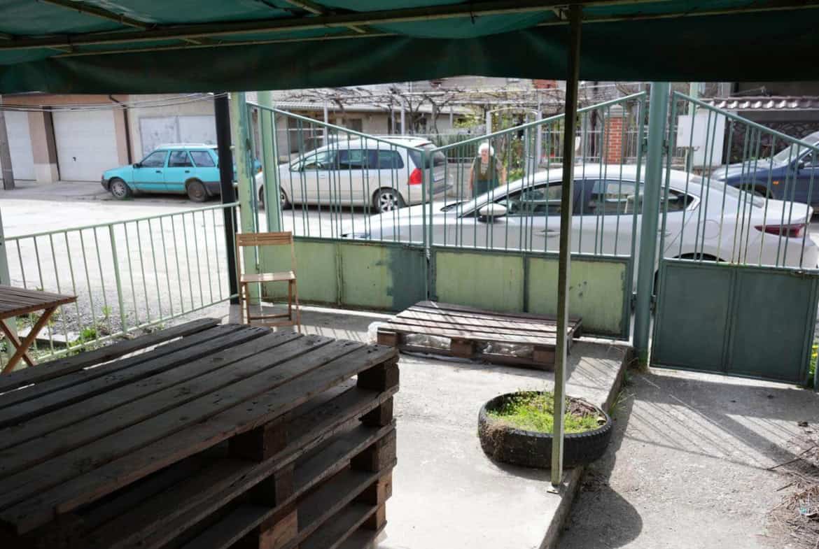 Веранда с дървени палети и стари столове, гледаща към улицата и паркирани автомобили.