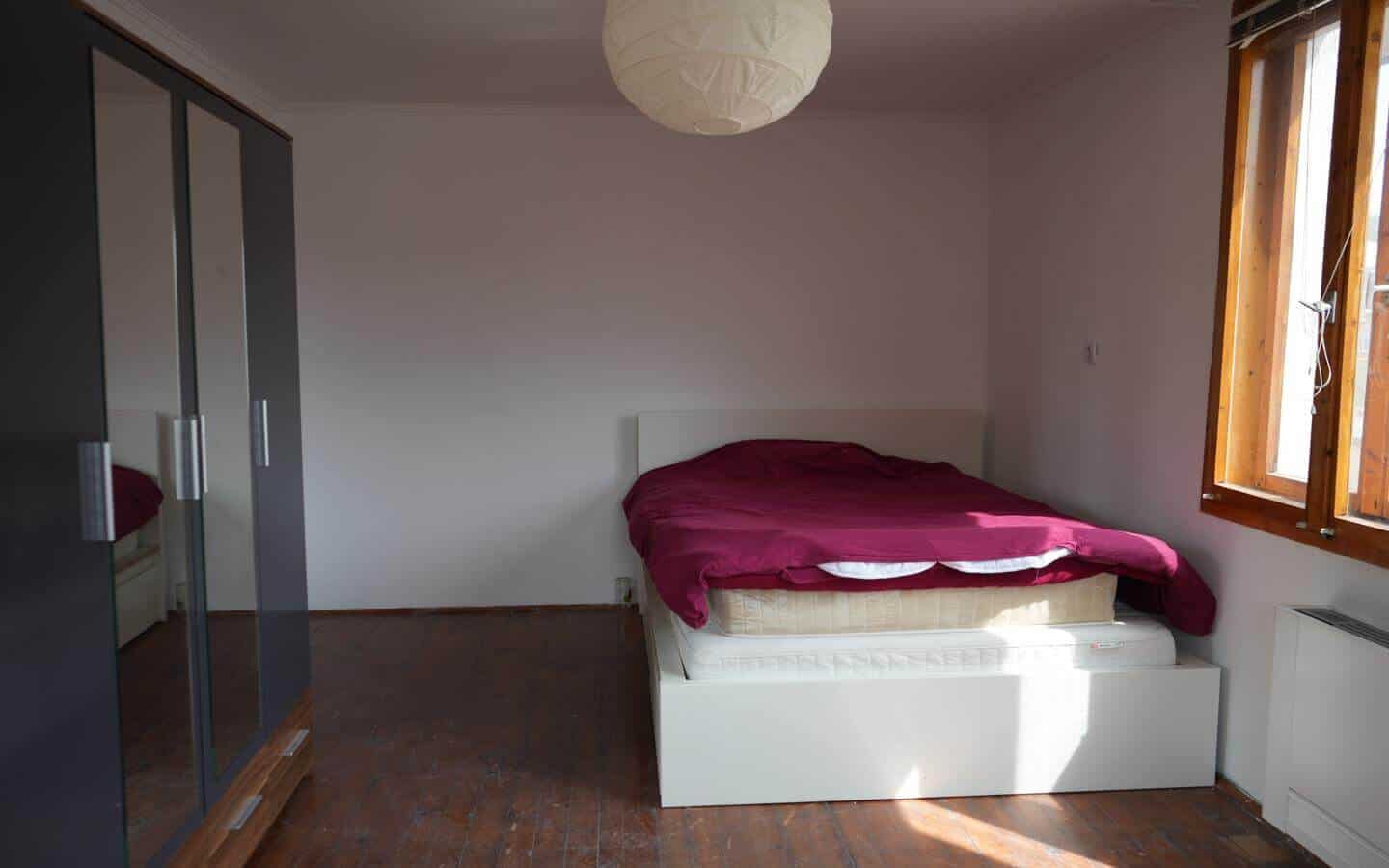 kushta-za-prodajba-kostenec-veranda-6 Спалня с двойно легло, облечено в тъмно червено чаршаф и голям гардероб.