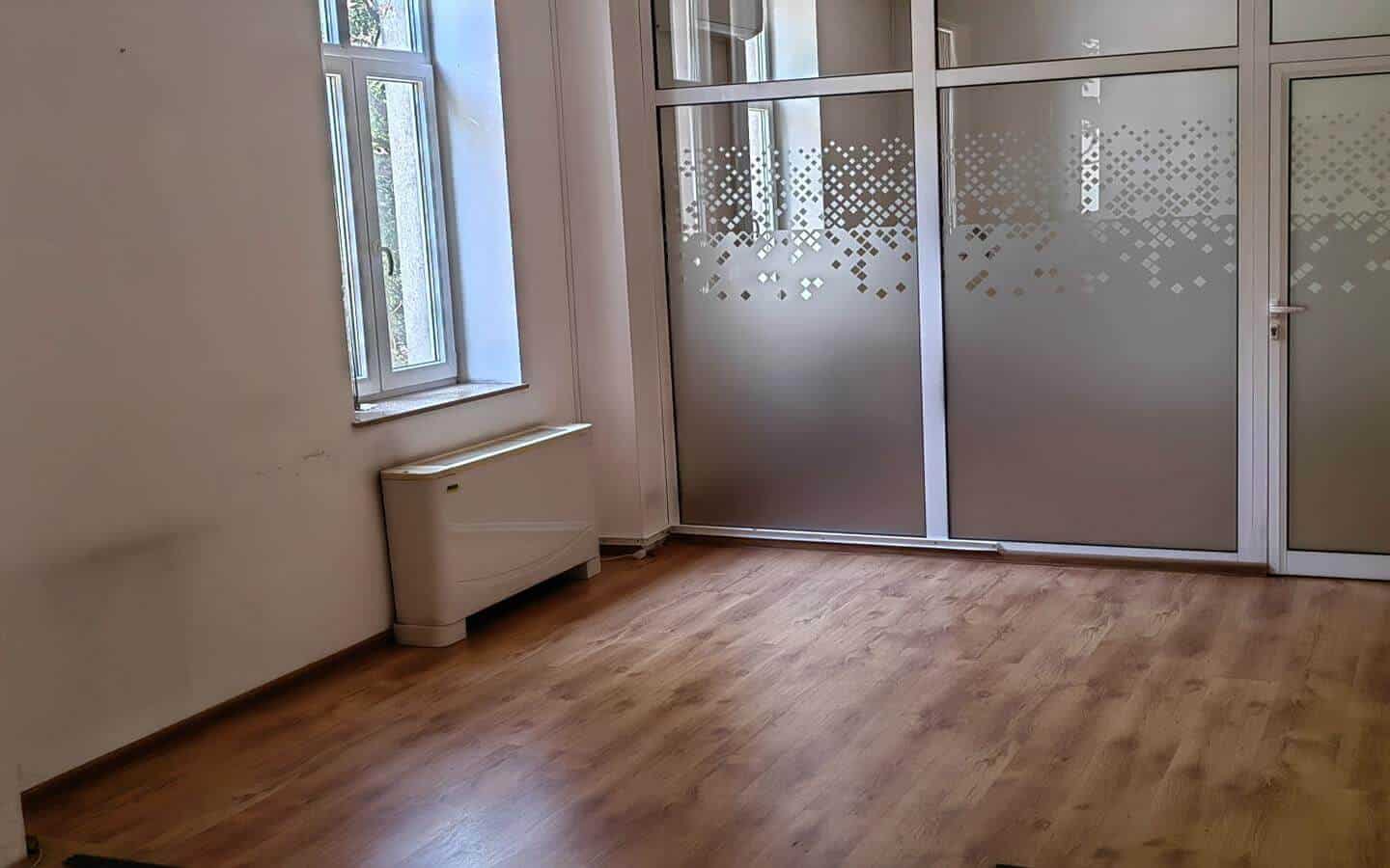 Празна стая с голям прозорец и стъклена врата с матово покритие от офис под наем в София до Централни хали.