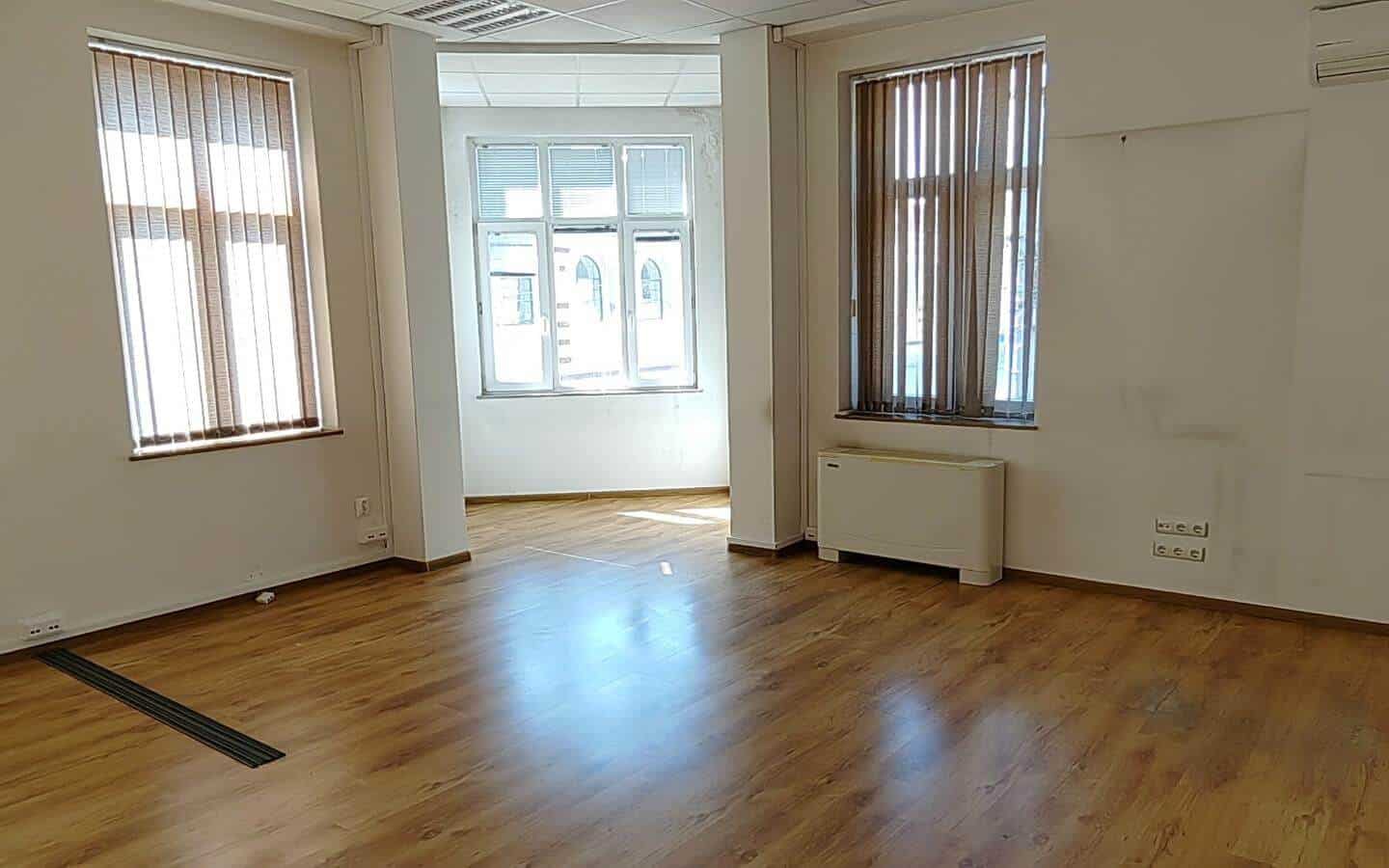 Празна стая с дървен под и три прозореца от офис под наем в София до Централни хали.