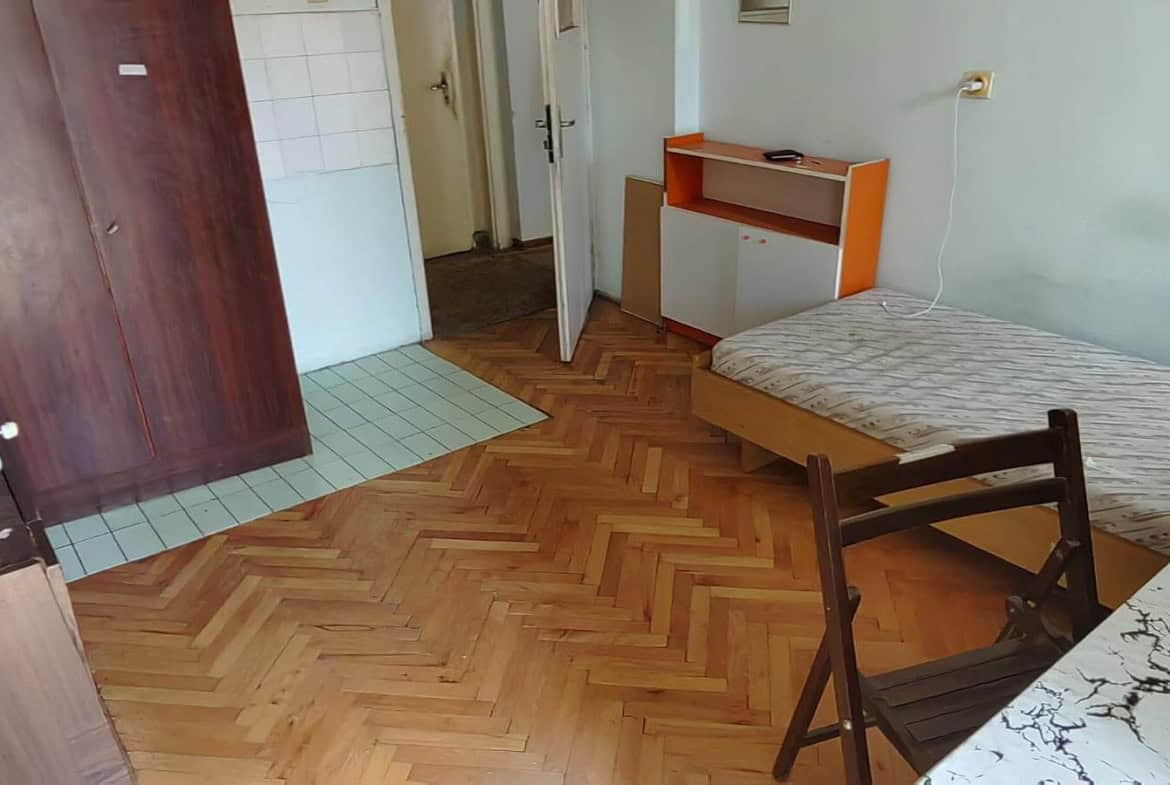 Стая с легло, маса и голям шкаф, с отворена врата към коридора.