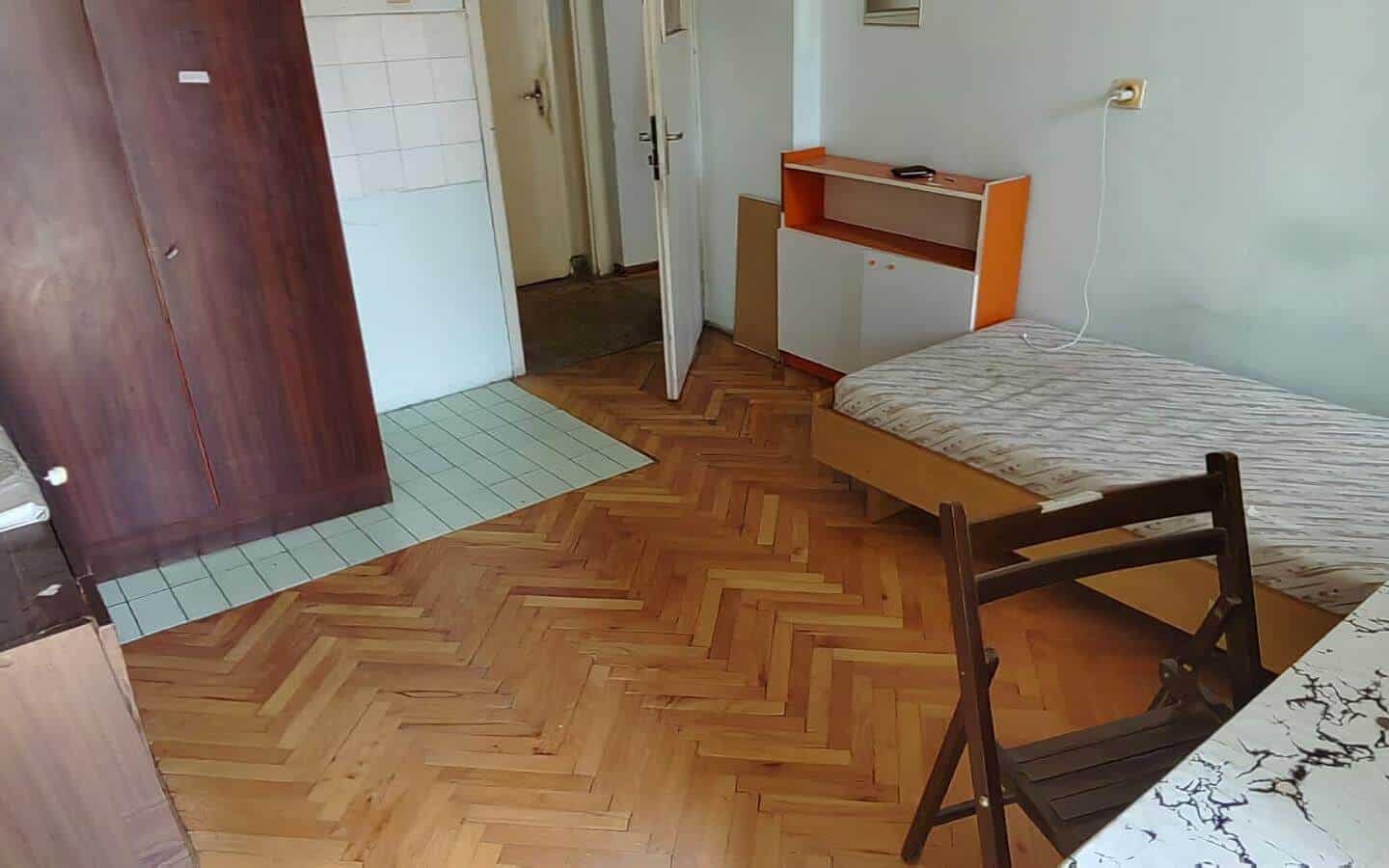 Стая с легло, маса и голям шкаф, с отворена врата към коридора.