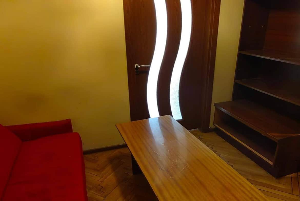 Хол с червен диван, дървена маса и шкаф, с врата с извита форма.