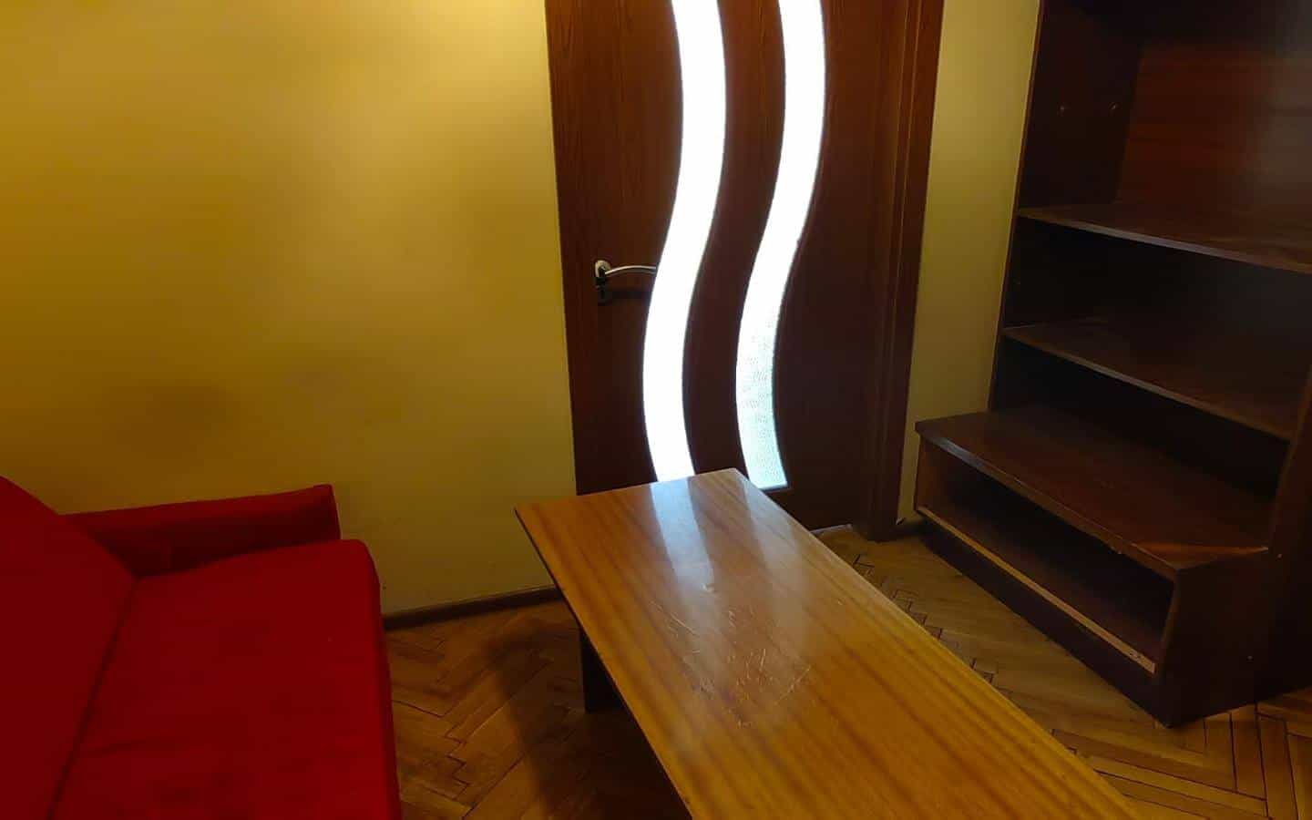 Хол с червен диван, дървена маса и шкаф, с врата с извита форма.