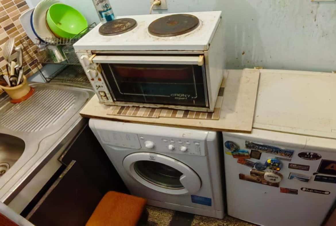 Малка кухня с мивка, пералня и малка печка върху хладилник.