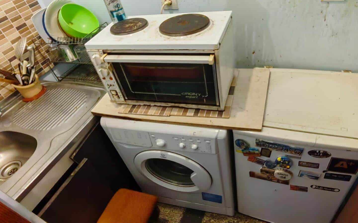 Малка кухня с мивка, пералня и малка печка върху хладилник.