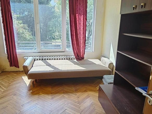 Стая с разтегателен диван, голям прозорец с червени пердета и шкаф.