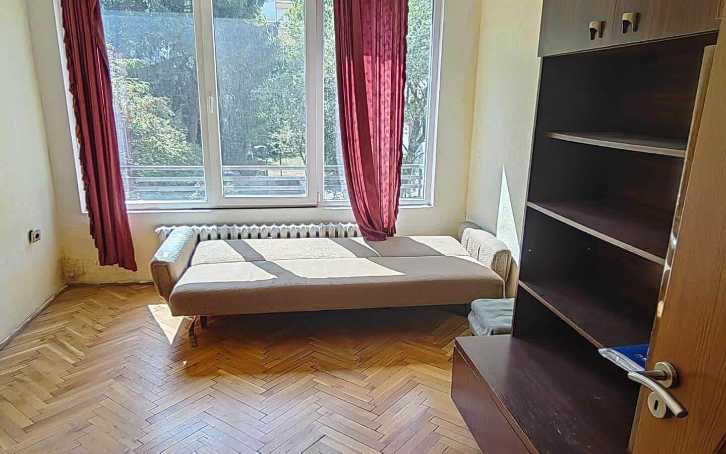 Стая с разтегателен диван, голям прозорец с червени пердета и шкаф.