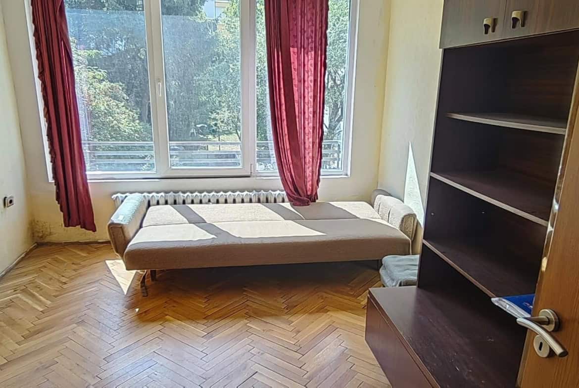 A room with a sofa bed, large window with red curtains, and a bookshelf.