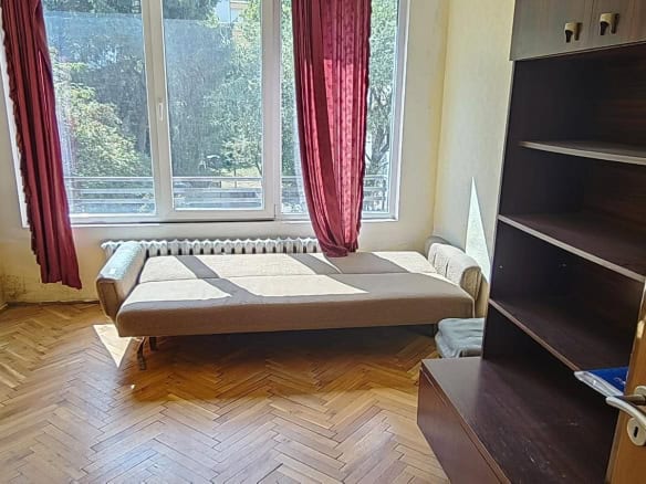 A room with a sofa bed, large window with red curtains, and a bookshelf.