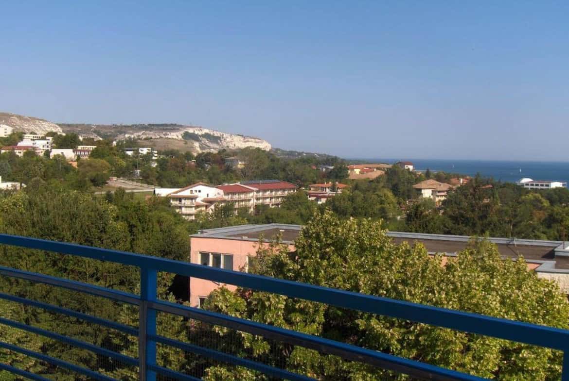 Панорамен изглед от балкона към крайбрежния пейзаж, с разположени ниско край морето къщи и високи планини в далечината.