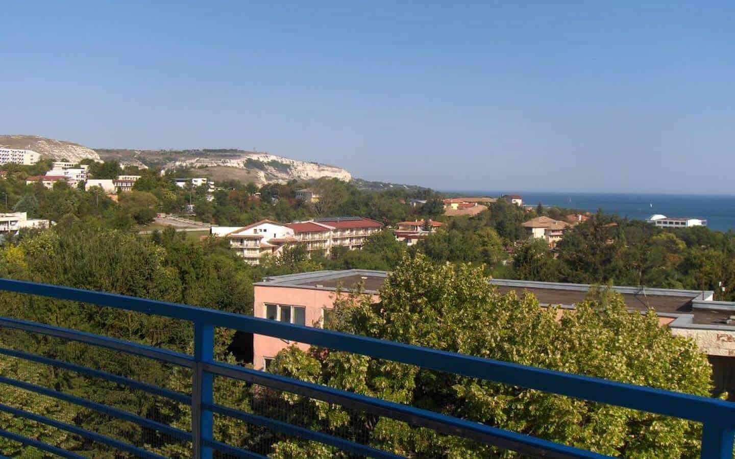 Панорамен изглед от балкона към крайбрежния пейзаж, с разположени ниско край морето къщи и високи планини в далечината.