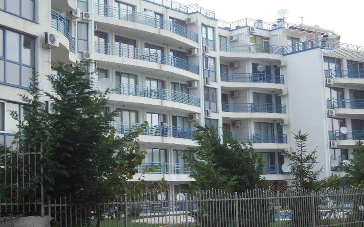 Модерен жилищен блок със светли фасади и сини балкони, заобиколен от зелени дървета и ограден с метална ограда.