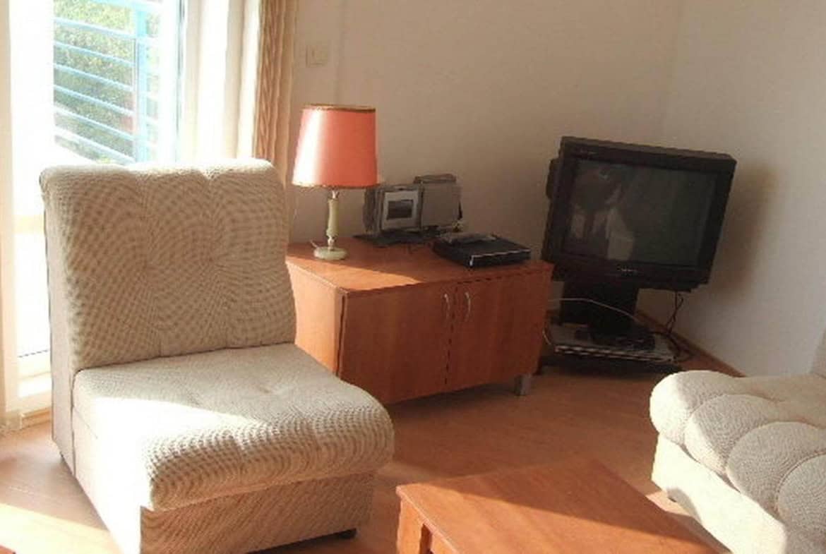 Светла всекидневна стая с два текстилни фотьойла и дървено бюро с лампа, до прозорец с изглед към дървета.