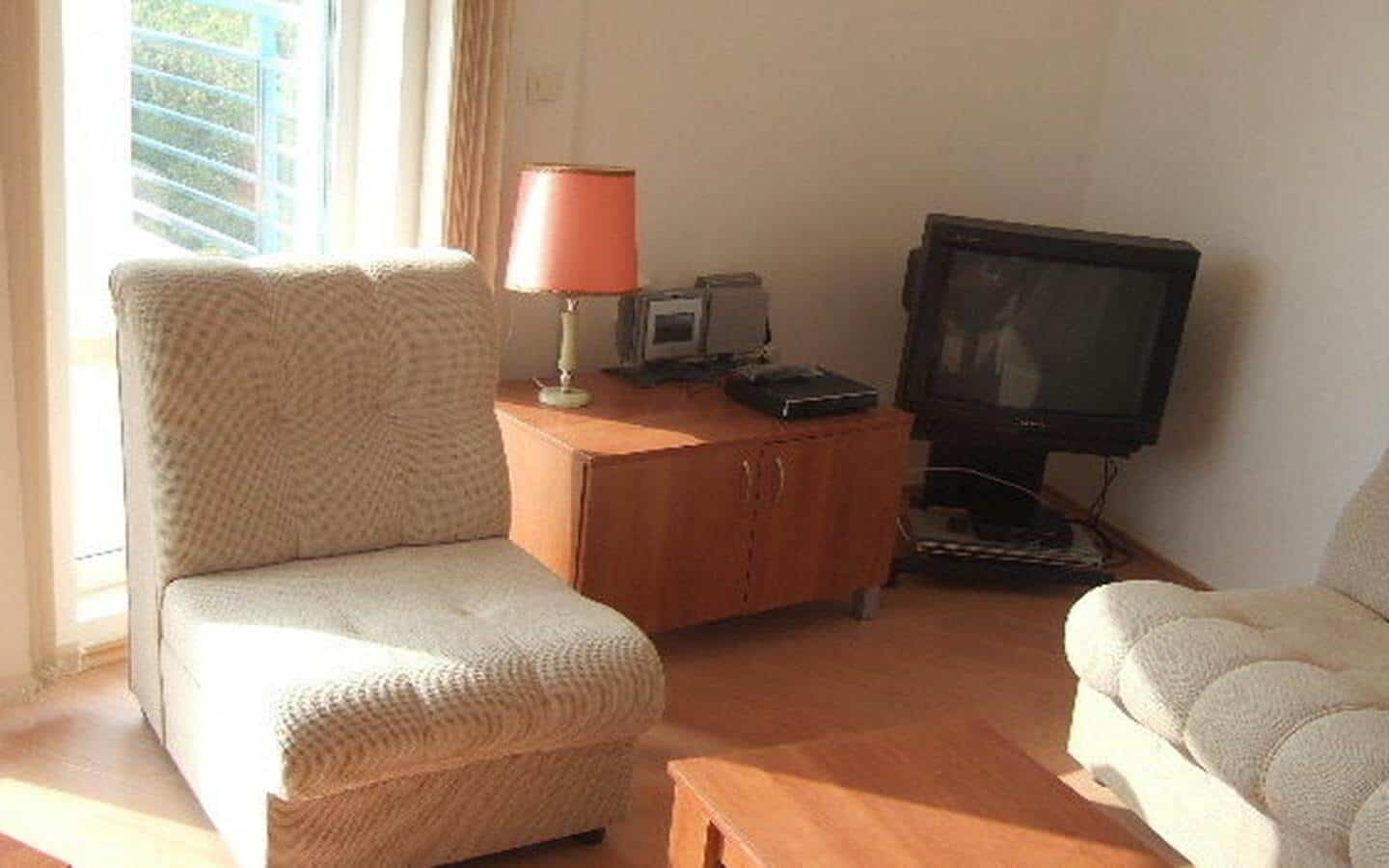 Светла всекидневна стая с два текстилни фотьойла и дървено бюро с лампа, до прозорец с изглед към дървета.