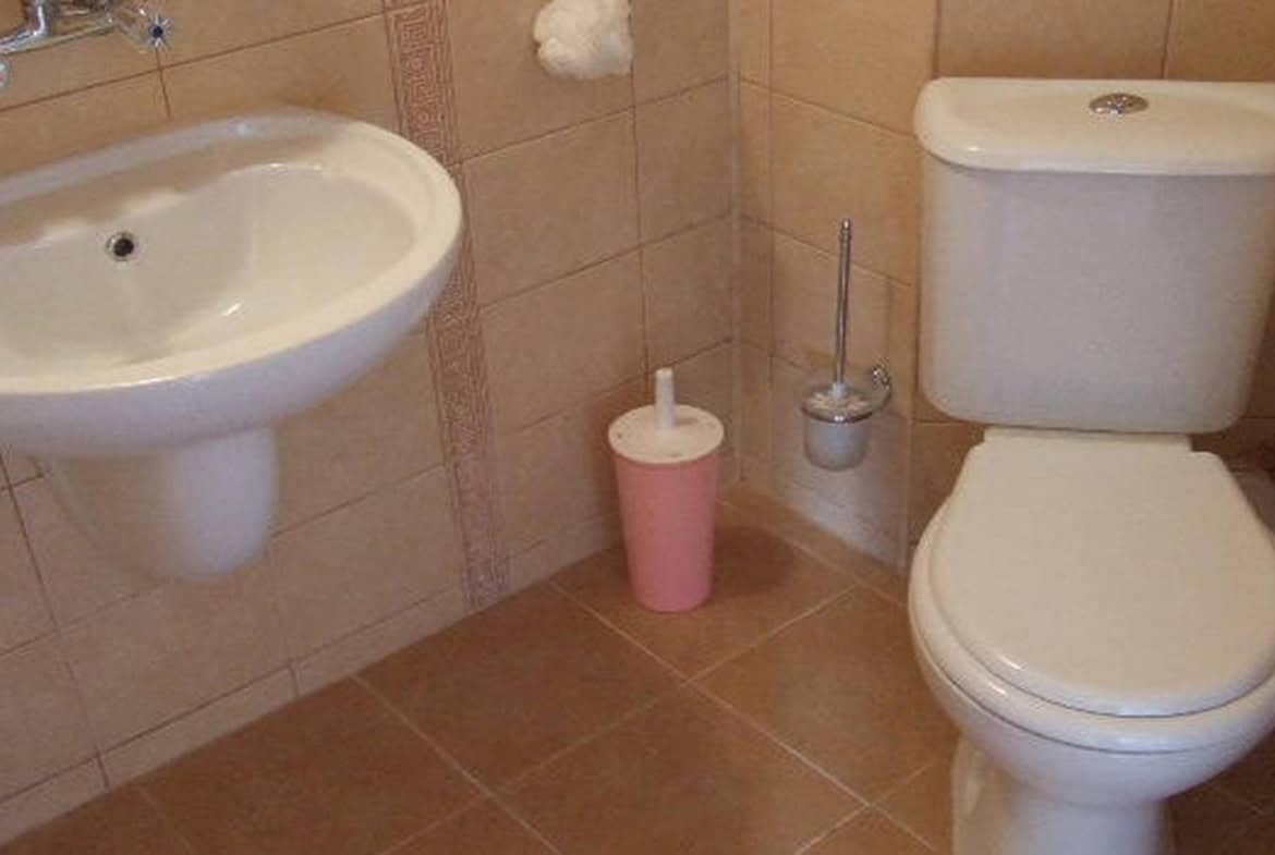 Баня със светло розови аксесоари, включваща тоалетна и умивалник, на фона на тъмнокафяви плочки.