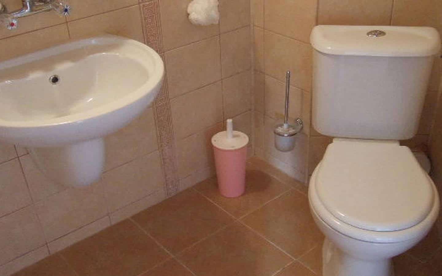 Баня със светло розови аксесоари, включваща тоалетна и умивалник, на фона на тъмнокафяви плочки.