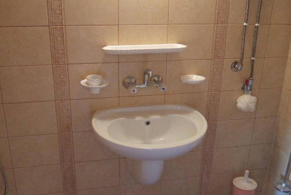 Умивалник с хромирани кранове и огледало, разположени на стена, облицована с кафяви плочки.