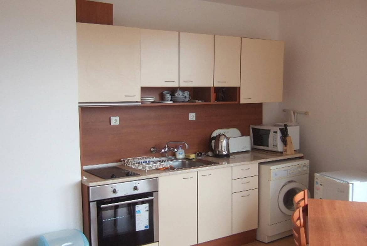 Малка кухня с бежови шкафове и дървени акценти, включваща печка, мивка и пералня, подредена в компактно пространство.