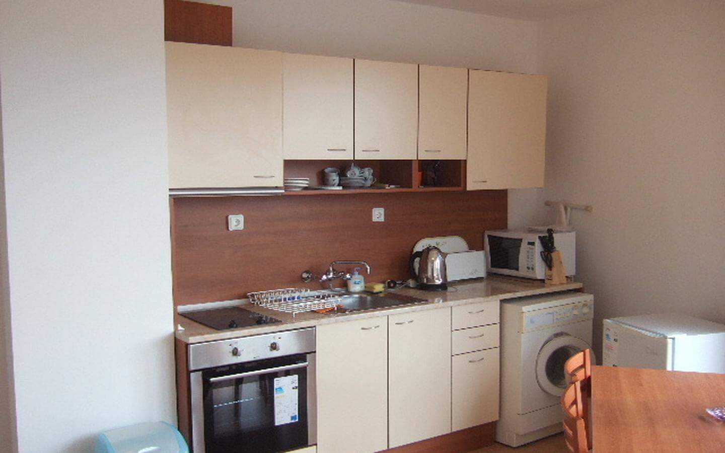 Малка кухня с бежови шкафове и дървени акценти, включваща печка, мивка и пералня, подредена в компактно пространство.