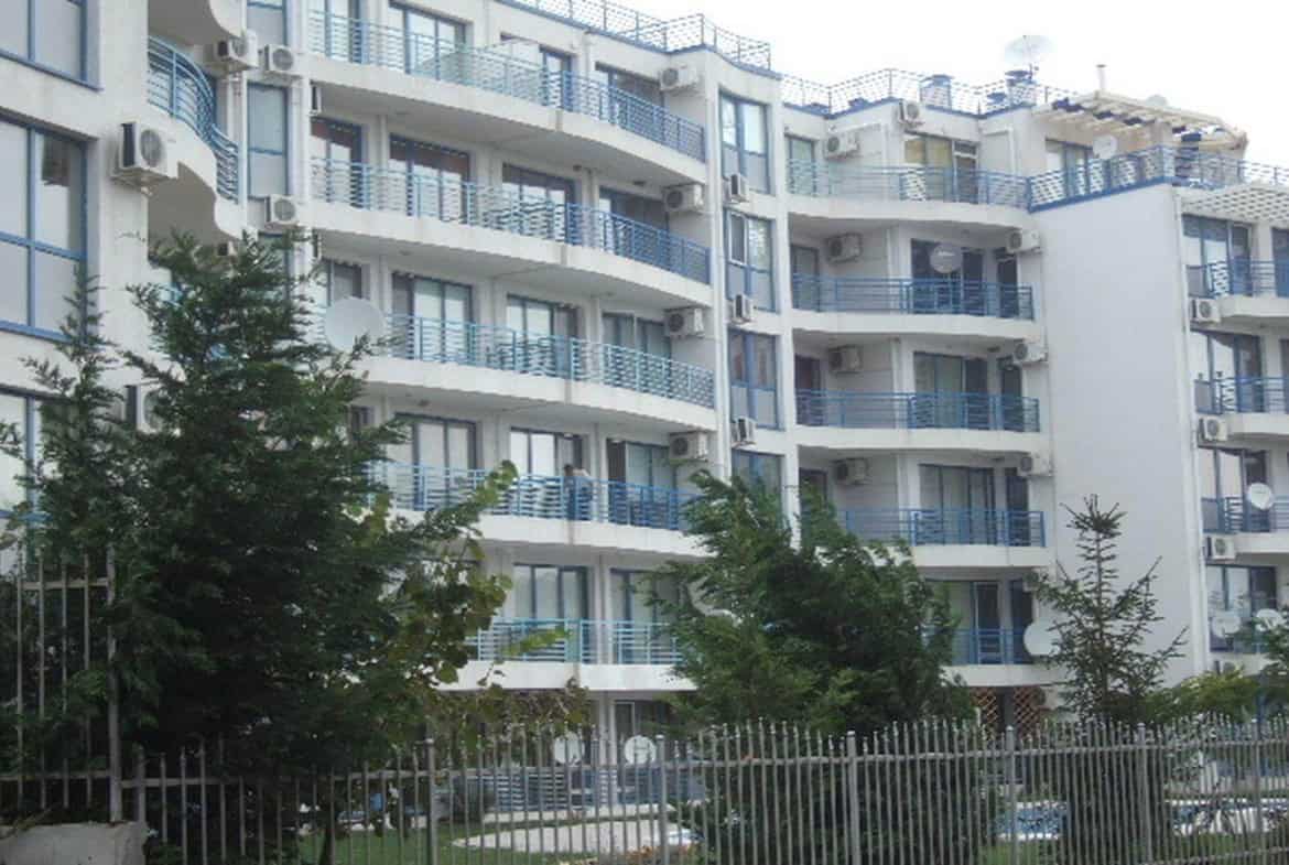 A modern residential building with light-colored facades and blue balconies, nestled among green trees and enclosed by a metal fence.