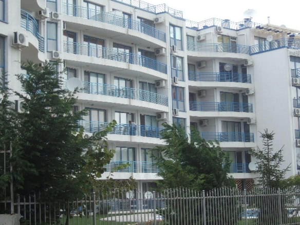 A modern residential building with light-colored facades and blue balconies, nestled among green trees and enclosed by a metal fence.