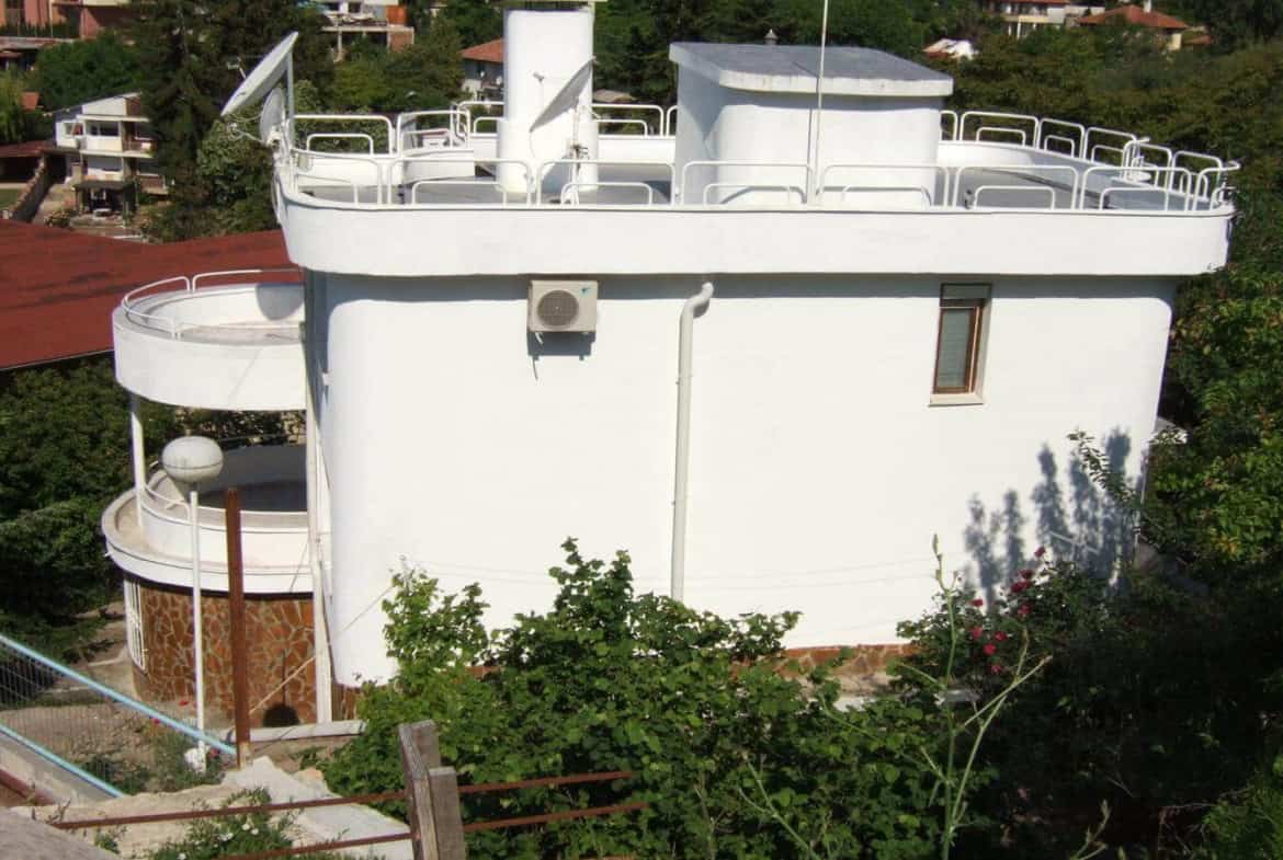 A peculiar white, round building with a flat roof, surrounded by white railings and equipped with several satellite dishes, set amidst green bushes.