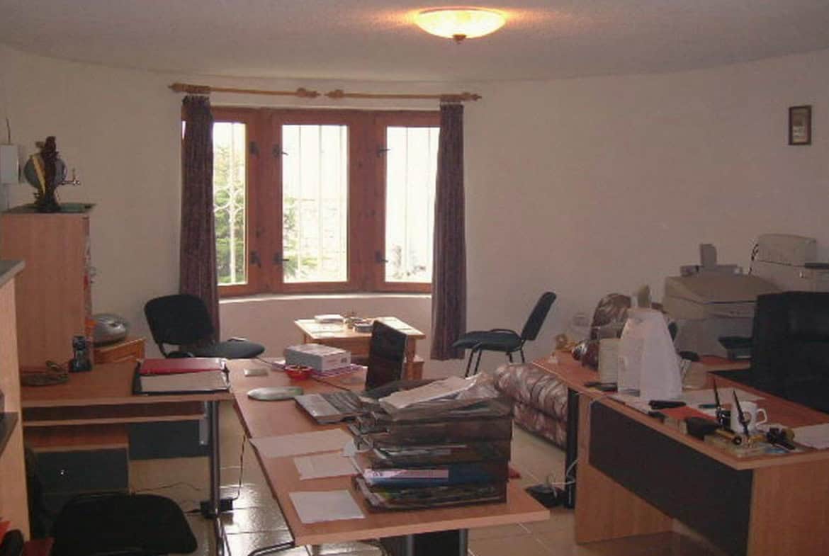 работно бюро в кабинет, пълно с книги, документи и офис оборудване, подредено в светла стая с прозорец, който показва зеленина навън.