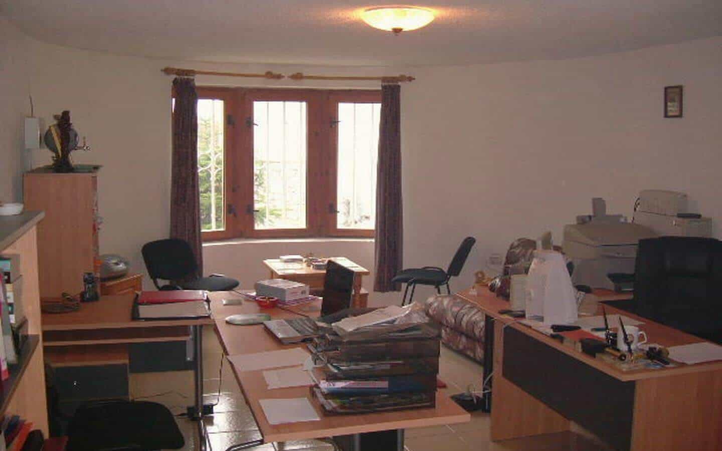 работно бюро в кабинет, пълно с книги, документи и офис оборудване, подредено в светла стая с прозорец, който показва зеленина навън.