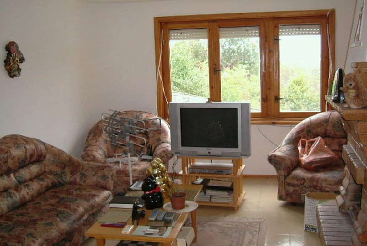 Уютен дневен тракт с два дивана и един фотьойл, покрити с цветни калъфки, пред телевизор, насочени към прозорец с изглед към листни дървета.