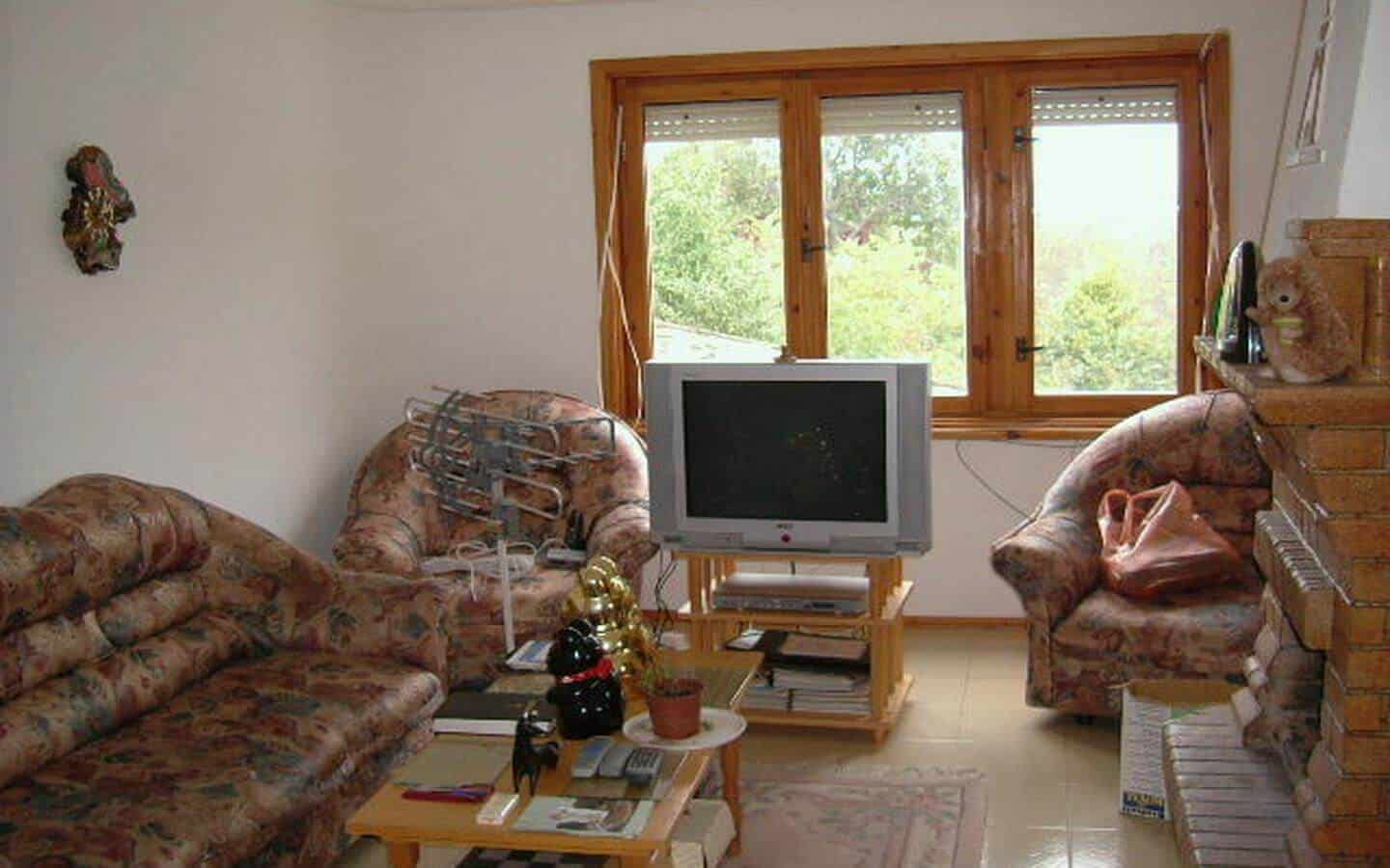 Уютен дневен тракт с два дивана и един фотьойл, покрити с цветни калъфки, пред телевизор, насочени към прозорец с изглед към листни дървета.