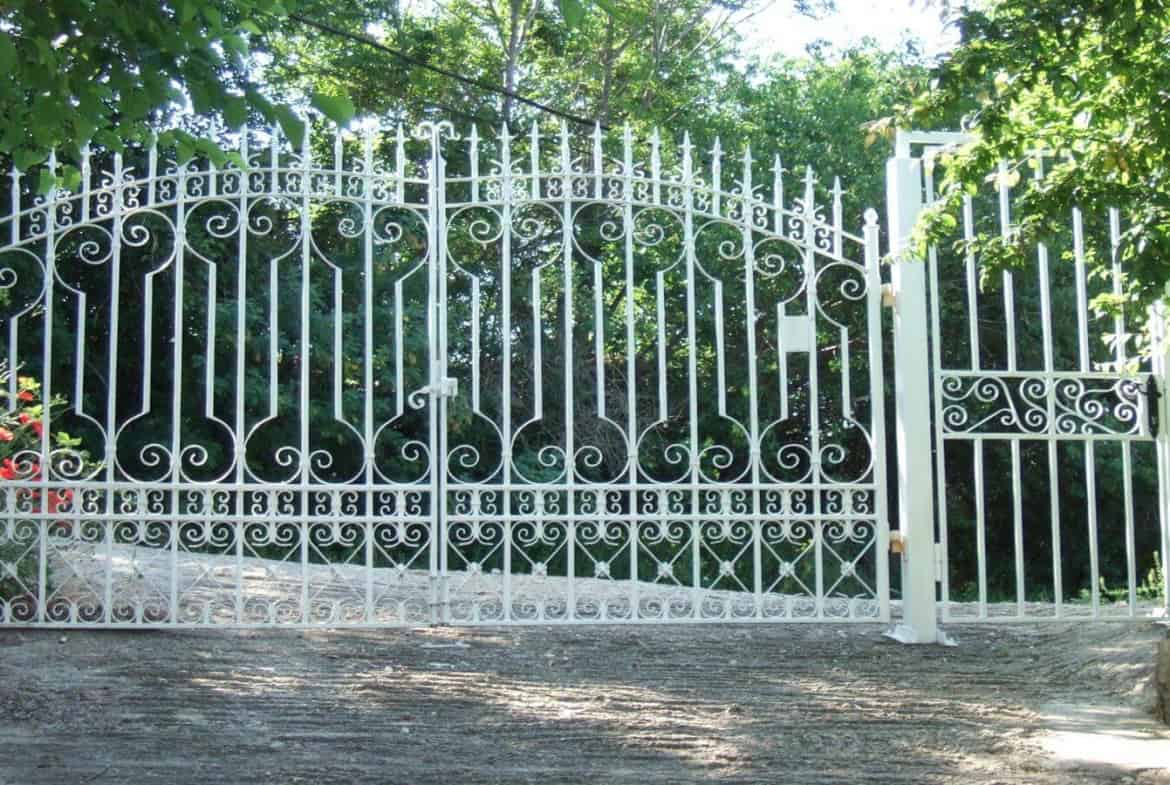 Елегантна бяла метална порта с изискани орнаменти и модели, отваряща се към пътека, заобиколена от зелени храсти и цветя.