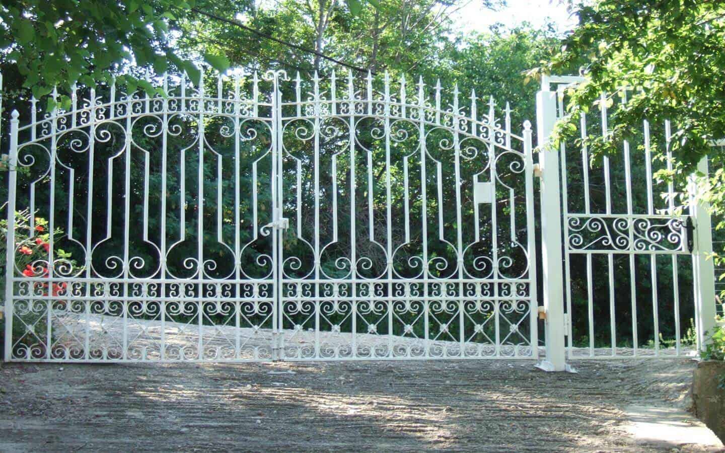 Елегантна бяла метална порта с изискани орнаменти и модели, отваряща се към пътека, заобиколена от зелени храсти и цветя.