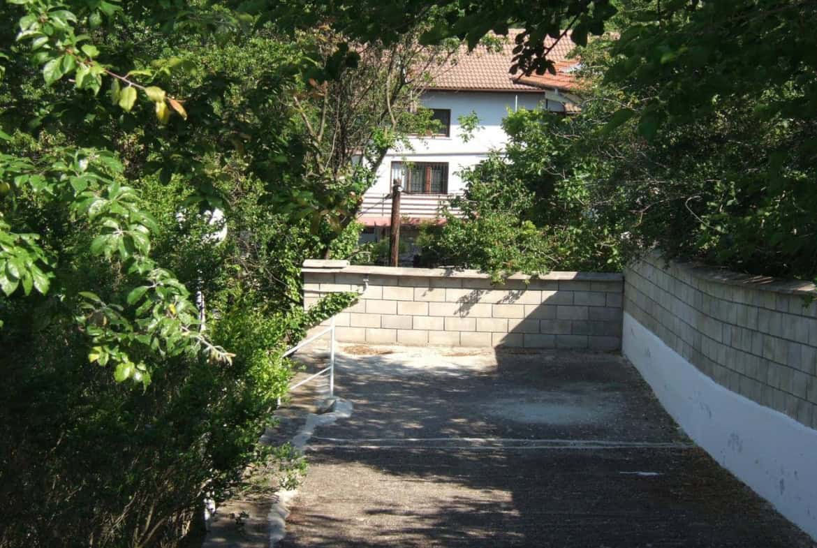 Снимка на алея вдясно от висока ограда, водеща към бяла къща, засенчена от листни дървета и храсти.