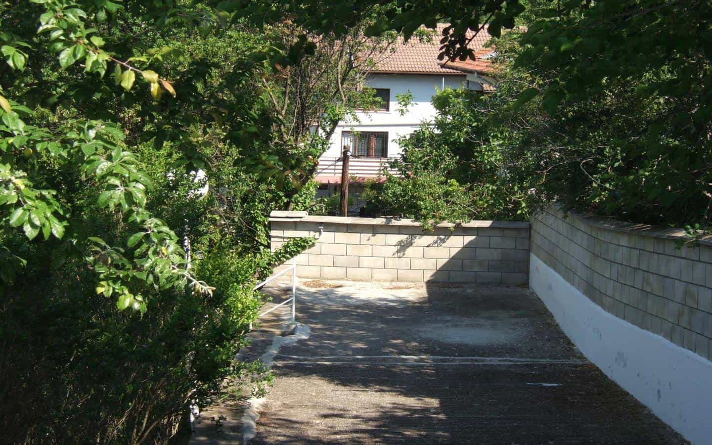 Снимка на алея вдясно от висока ограда, водеща към бяла къща, засенчена от листни дървета и храсти.