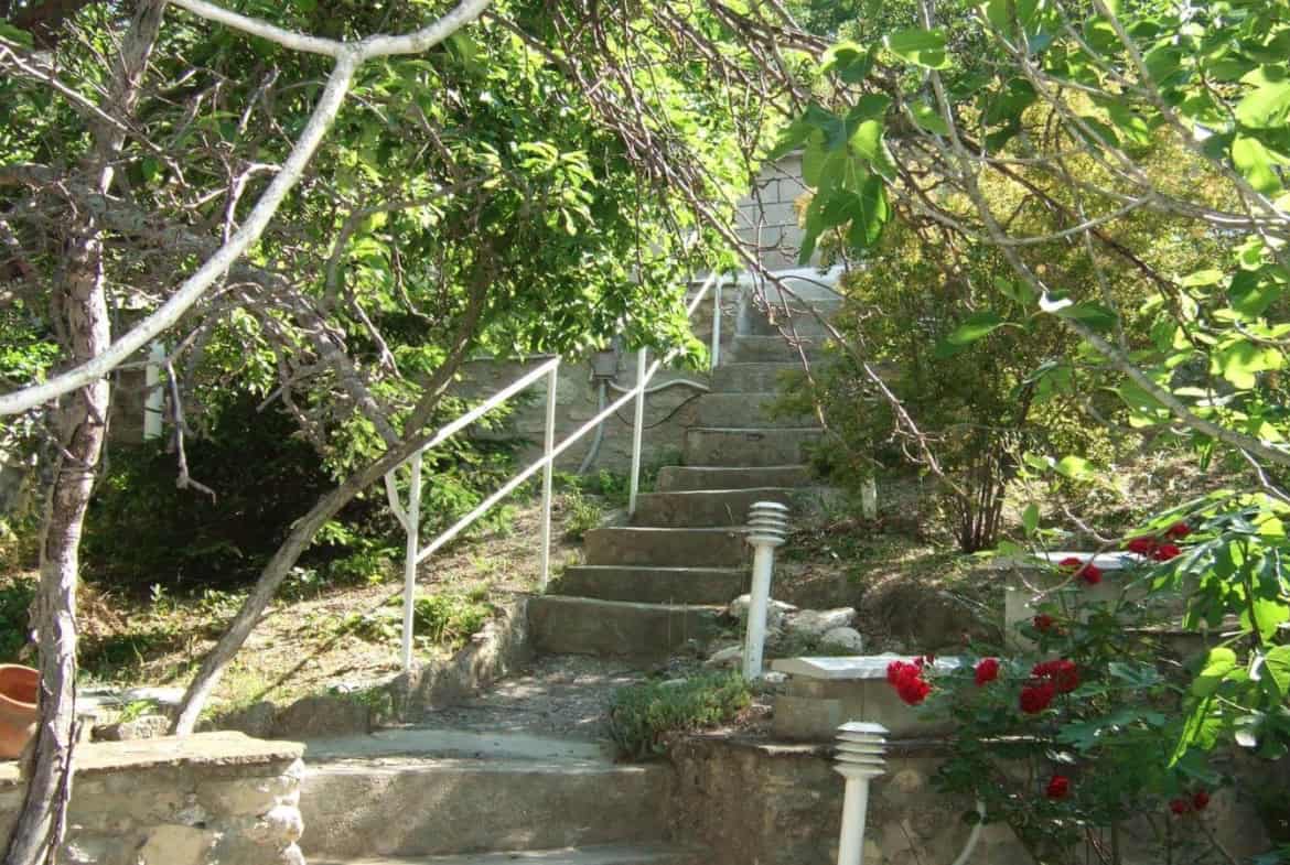Kаменнo стълбище, обграденo със зелени растения и цветя, водещo към светла сграда в края.