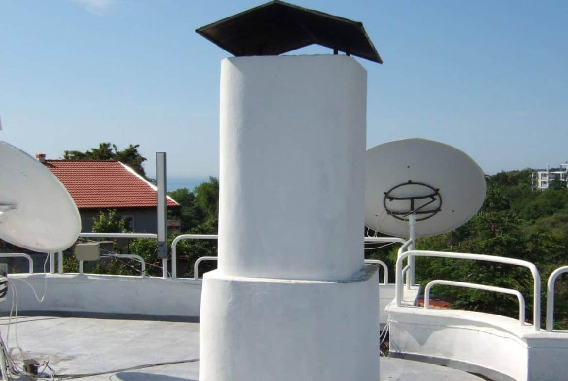 Голям бял комин с плосък покрив, заобиколен от сателитни антени на покрива на сграда с изглед към морския хоризонт.