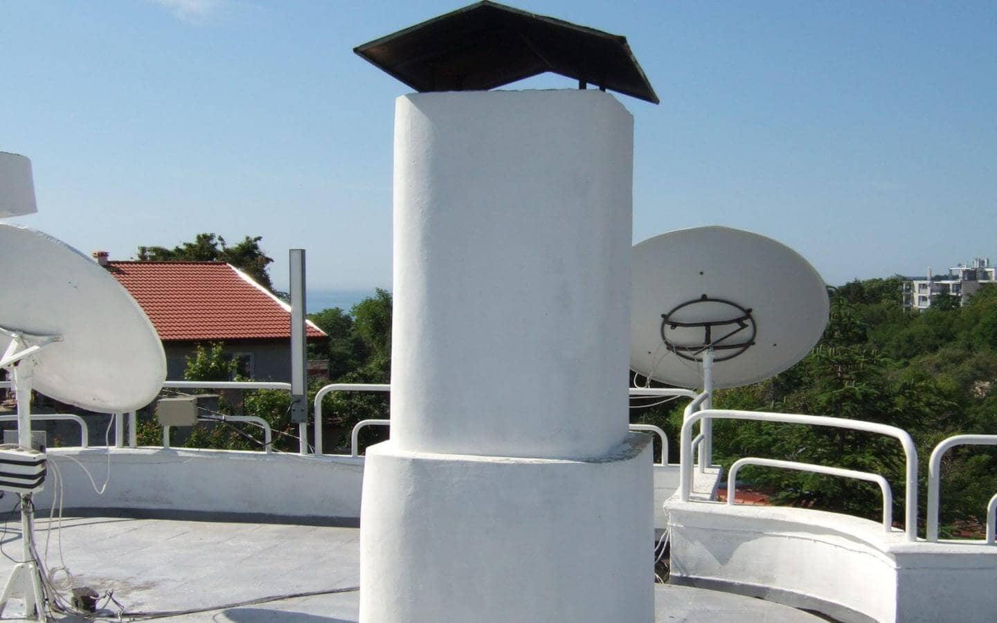 Голям бял комин с плосък покрив, заобиколен от сателитни антени на покрива на сграда с изглед към морския хоризонт.